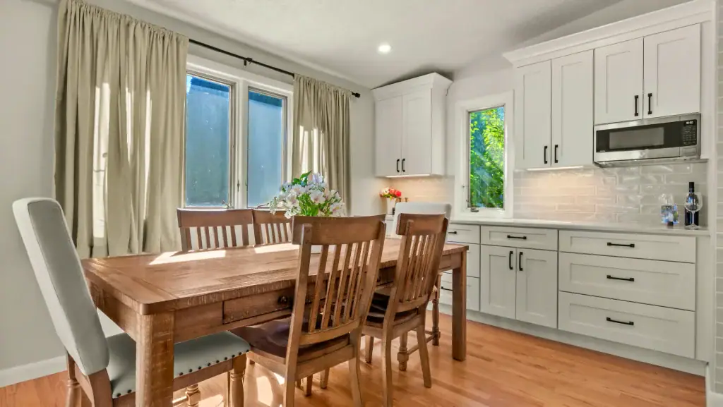 Wood dining table beside white kitchen cabinetry, quartz counters, and a window overlooking greenery.