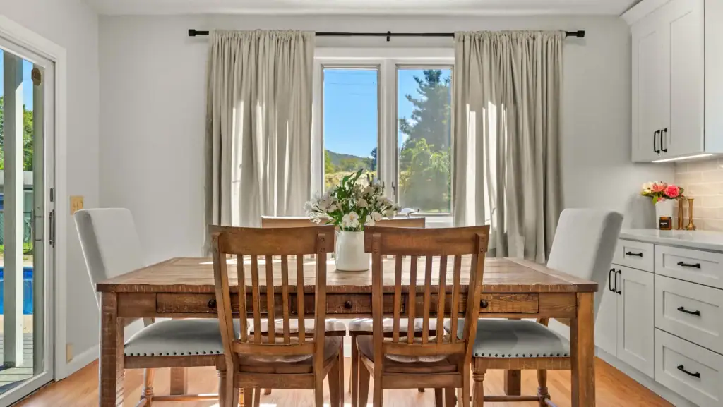 Sunlit dining nook with a wood table, upholstered bench, and white built-in kitchen cabinetry.