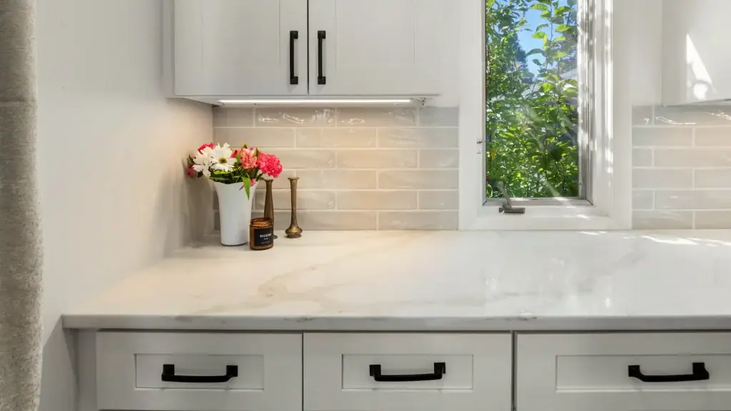 Quartz countertop, white lower cabinets, pale tile backsplash, and a narrow window with leafy views.